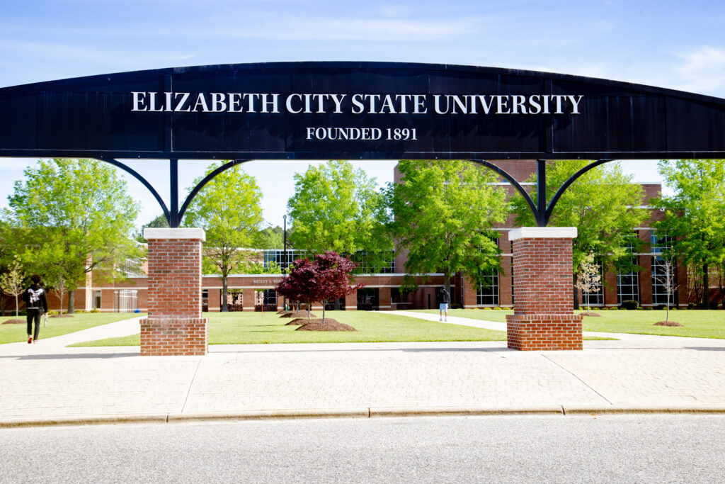 The ECSU arch displays the text “Elizabeth City State University, Founded 1891” at the entrance to a campus quad. Academic buildings, walkways, trees, and students are visible beyond the arch on a sunny day.