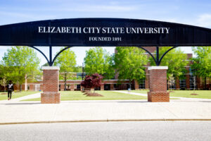 The ECSU arch displays the text “Elizabeth City State University, Founded 1891” at the entrance to a campus quad. Academic buildings, walkways, trees, and students are visible beyond the arch on a sunny day.
