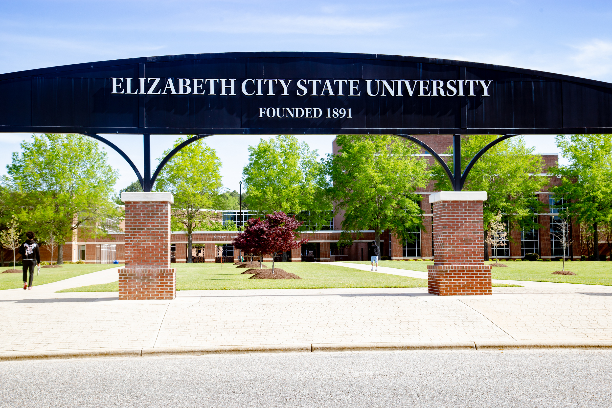 The ECSU arch displays the text “Elizabeth City State University, Founded 1891” at the entrance to a campus quad. Academic buildings, walkways, trees, and students are visible beyond the arch on a sunny day.
