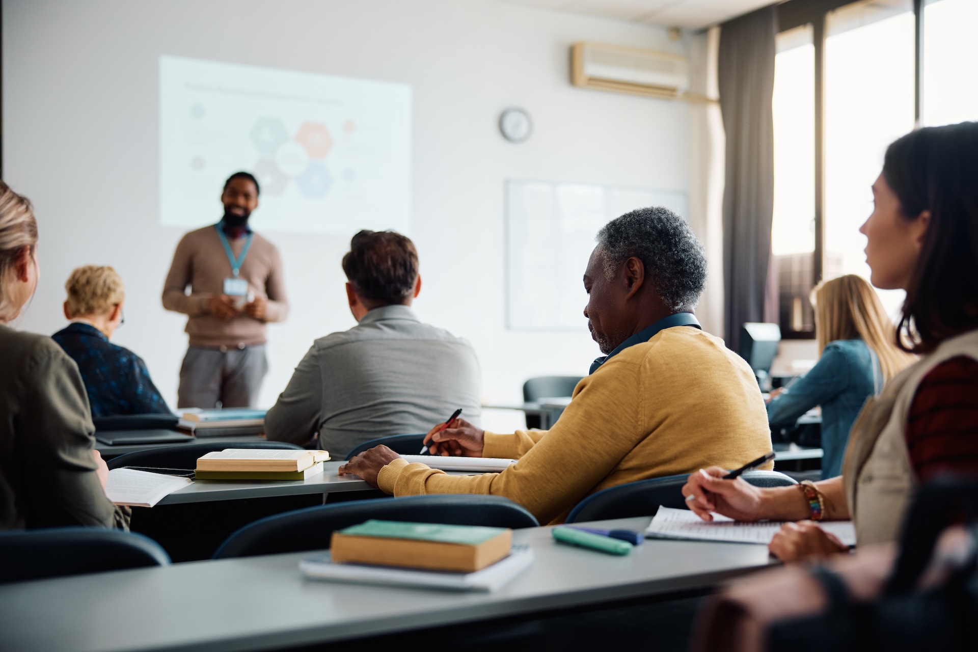Students of varied ages are seated at desks in a brightly lit classroom listening to a lecture. 