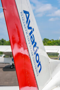 Close-up of the tail section of an ECSU Aviation training aircraft featuring Elizabeth City State University branding in blue lettering on a white surface with a red vertical stabilizer. Additional aircraft are visible in the background on the tarmac.