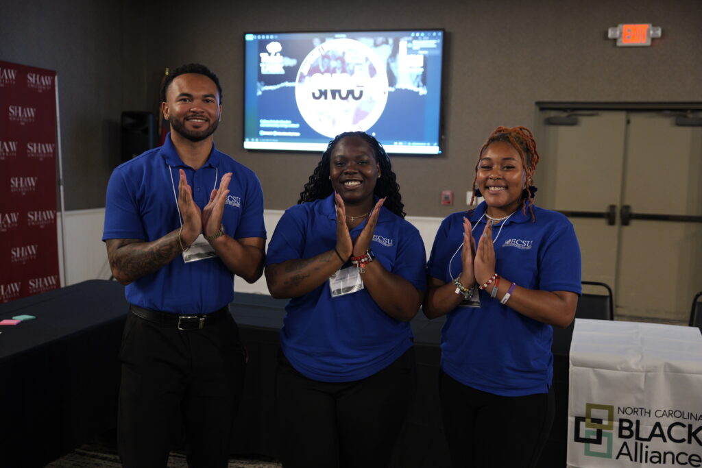 Three ECSU students wearing matching blue ECSU polo shirts stand together at a conference or networking event. A presentation screen and event signage are visible in the background.