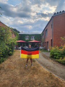 Student stands in a courtyard between ivy-covered brick buildings, holding a German flag. Outdoor seating with red umbrellas is visible in the background beneath a cloudy sky.