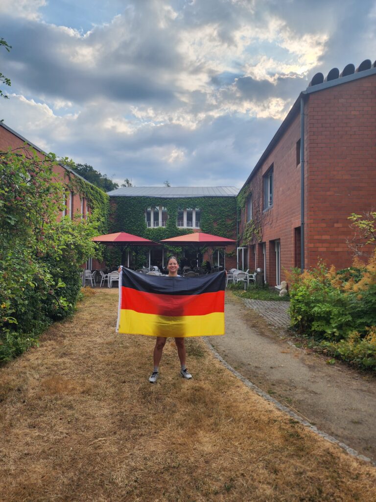 Student stands in a courtyard between ivy-covered brick buildings, holding a German flag. Outdoor seating with red umbrellas is visible in the background beneath a cloudy sky.